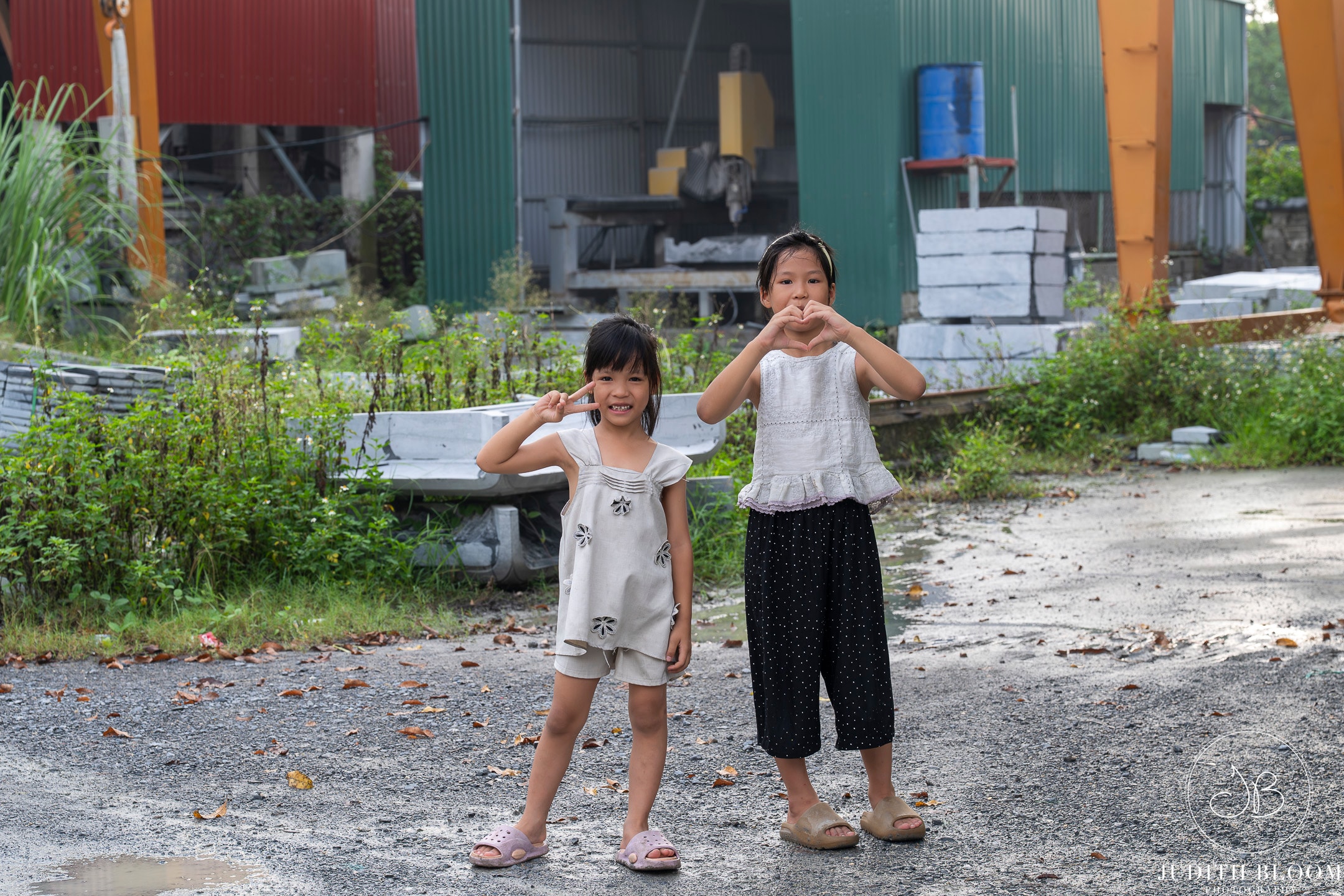 Two young Vietnamese girls pose playfully — one flashing a peace sign, the other a heart — in an industrial yard