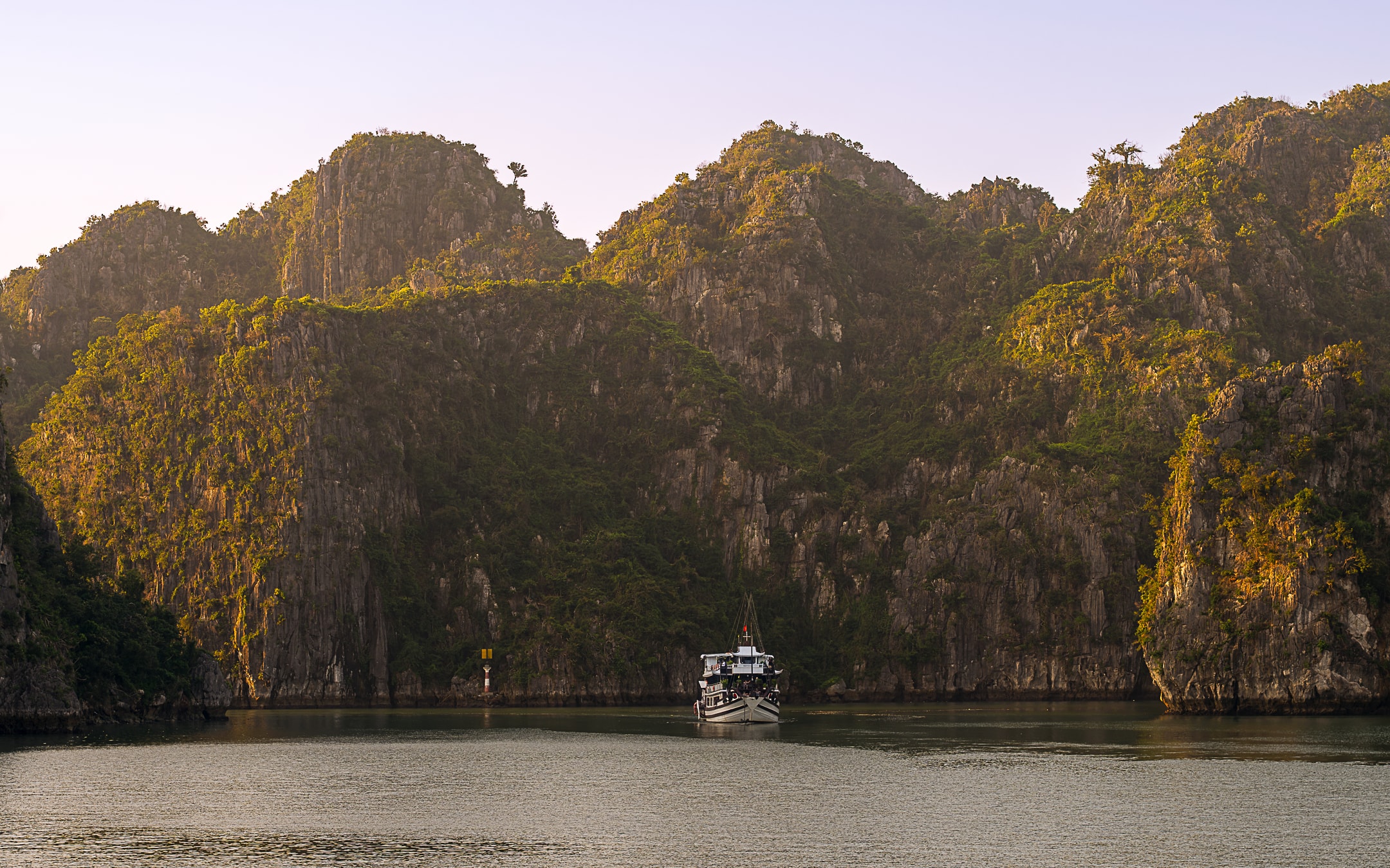 A traditional wooden cruise boat navigates between towering golden limestone karsts at dusk, Ha Long Bay