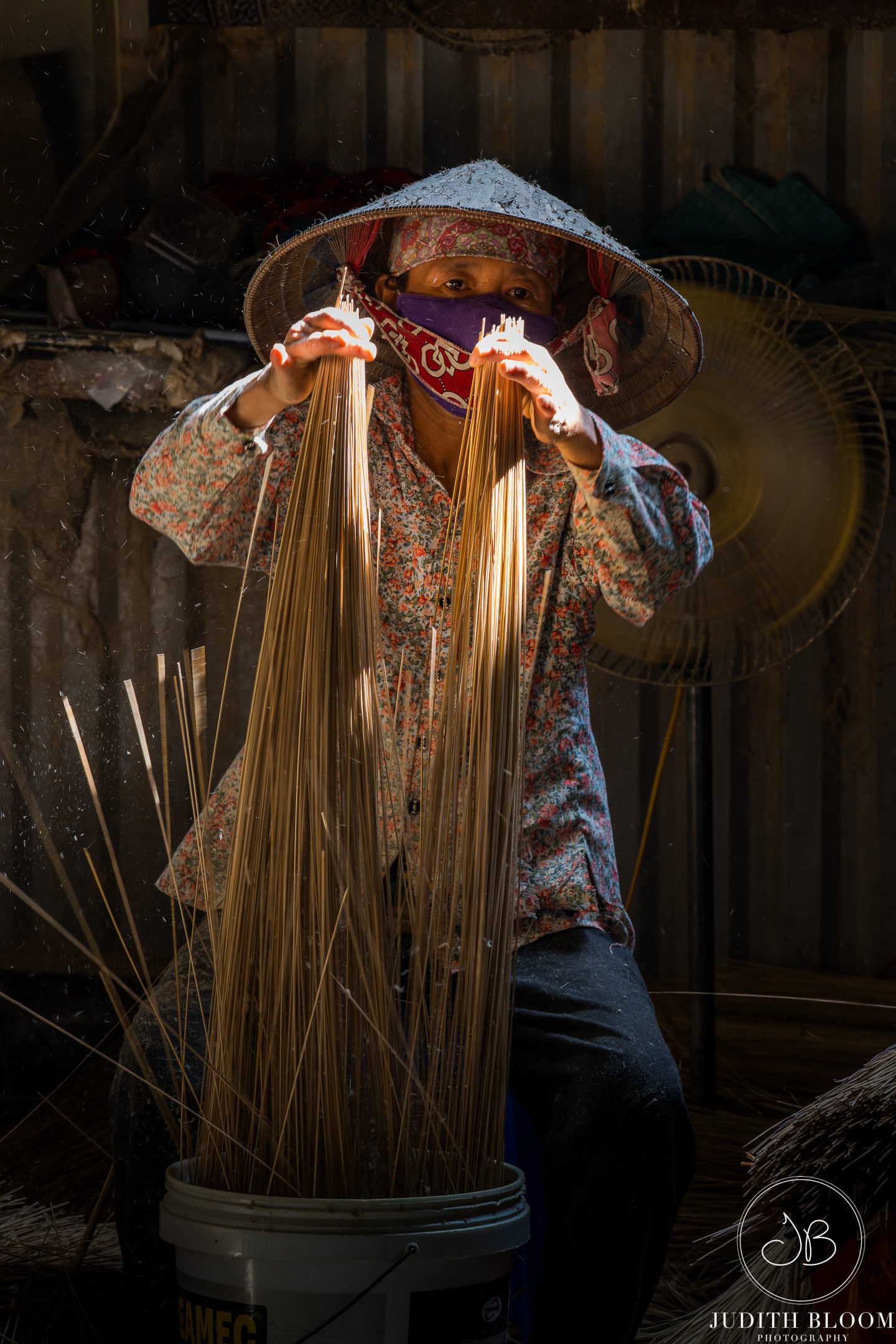 An incense maker in a conical hat works by dramatic light in a workshop