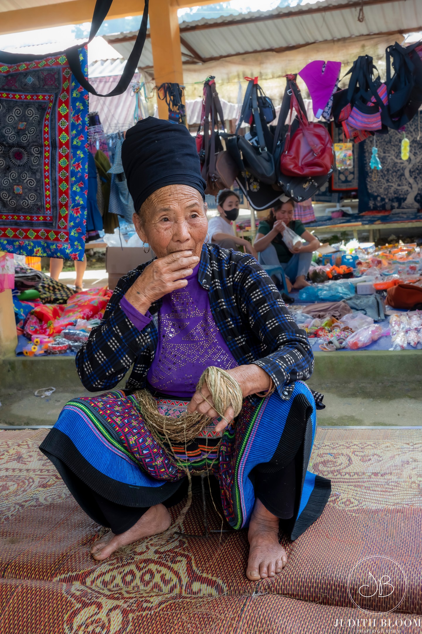 An elderly Hmong woman in traditional dress sits at a market stall, holding a bundle of natural fibre
