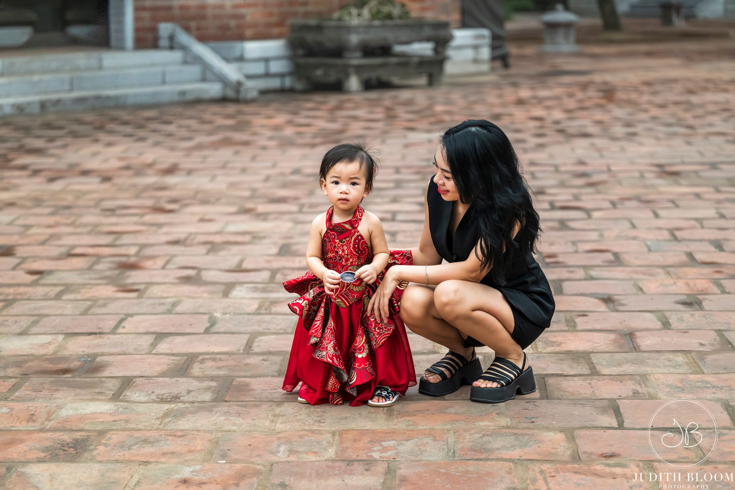 A mother kneels beside her young daughter in a red traditional Vietnamese dress on a brick courtyard
