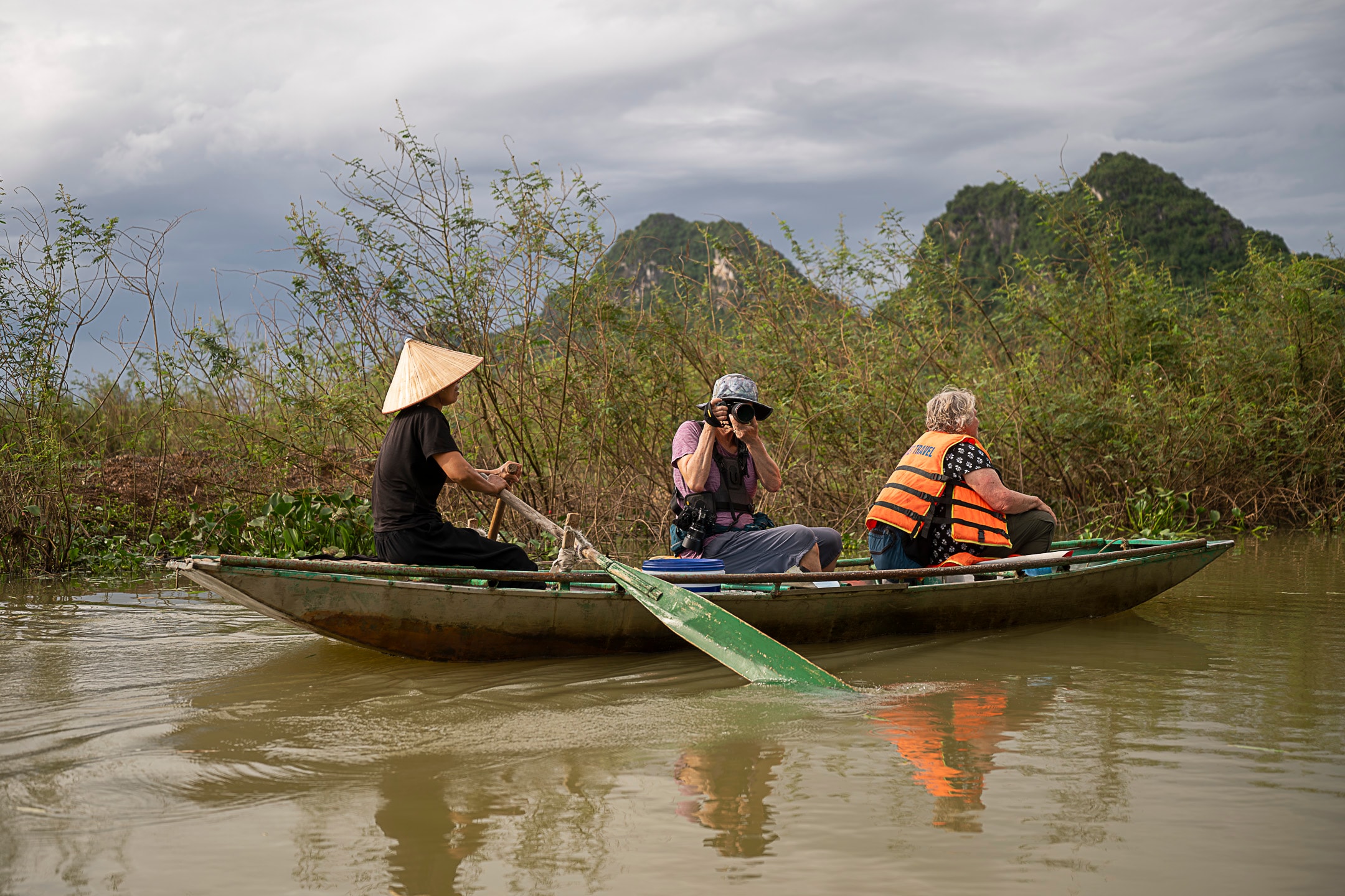 A local boatwoman rows two photographers through a narrow river channel beneath dramatic storm clouds