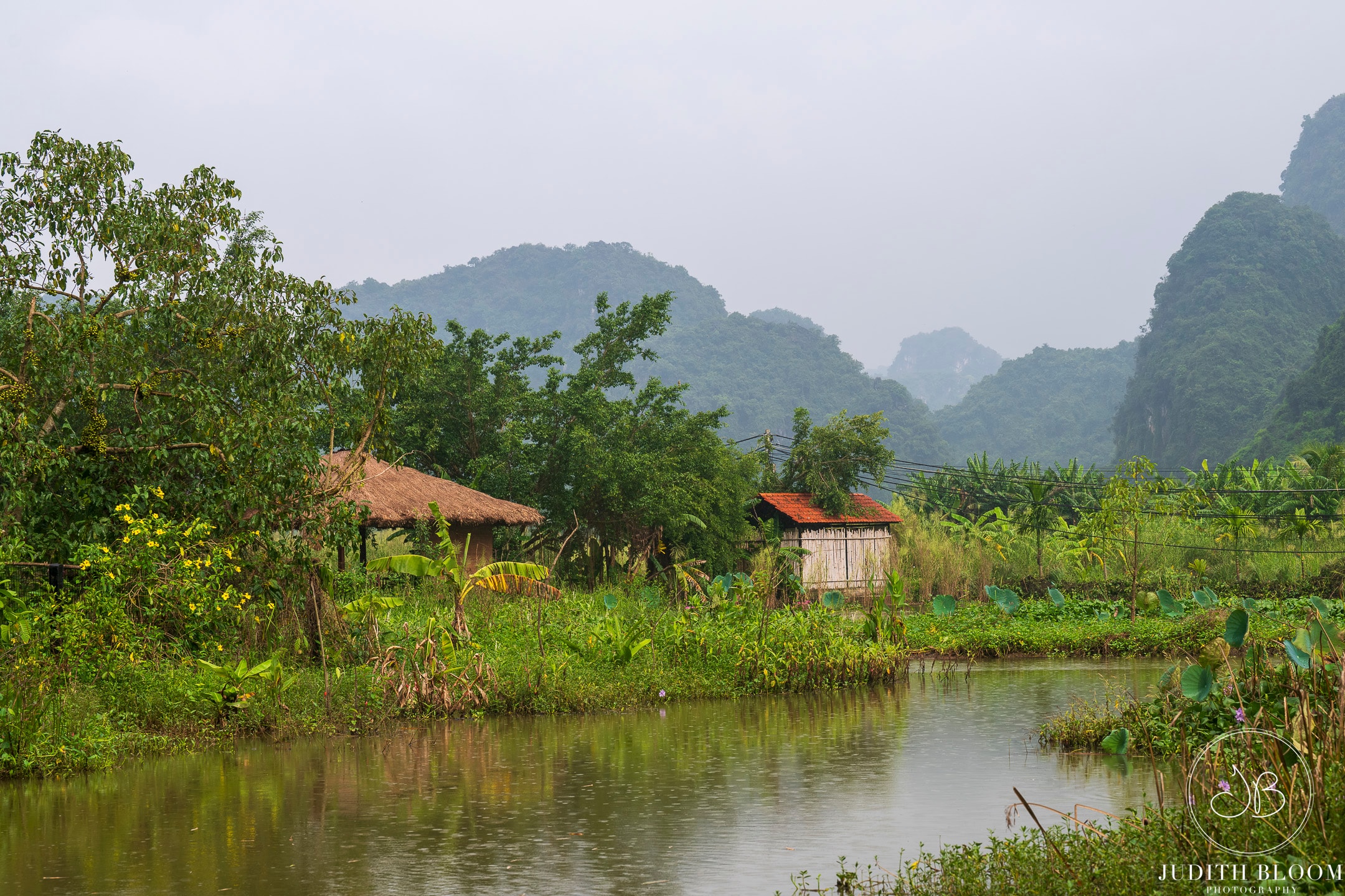 Misty karst mountains frame a quiet riverside village with thatched and tiled rooftops, Tam Coc