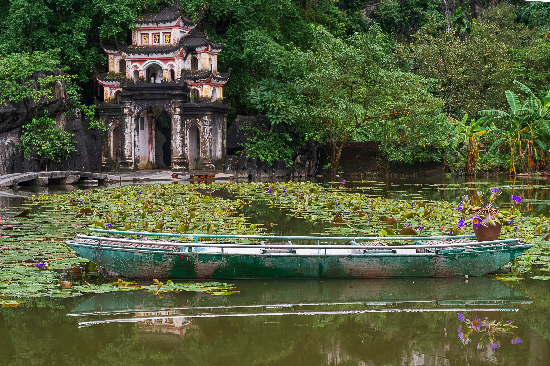 A weathered wooden boat rests among water lilies before an ornate temple gateway, Ninh Binh