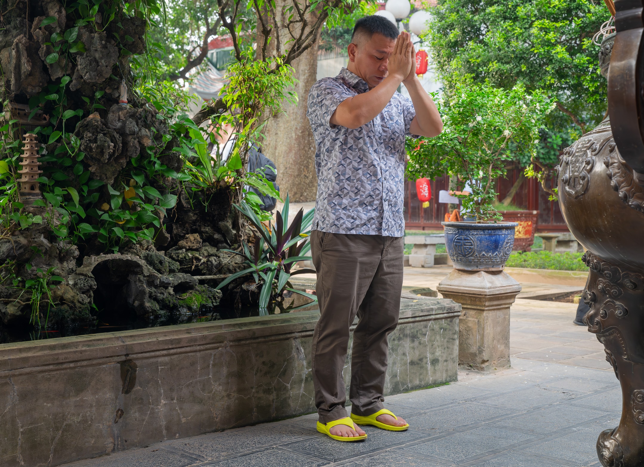 A man in quiet prayer at a temple courtyard surrounded by lush greenery and bronze vessels