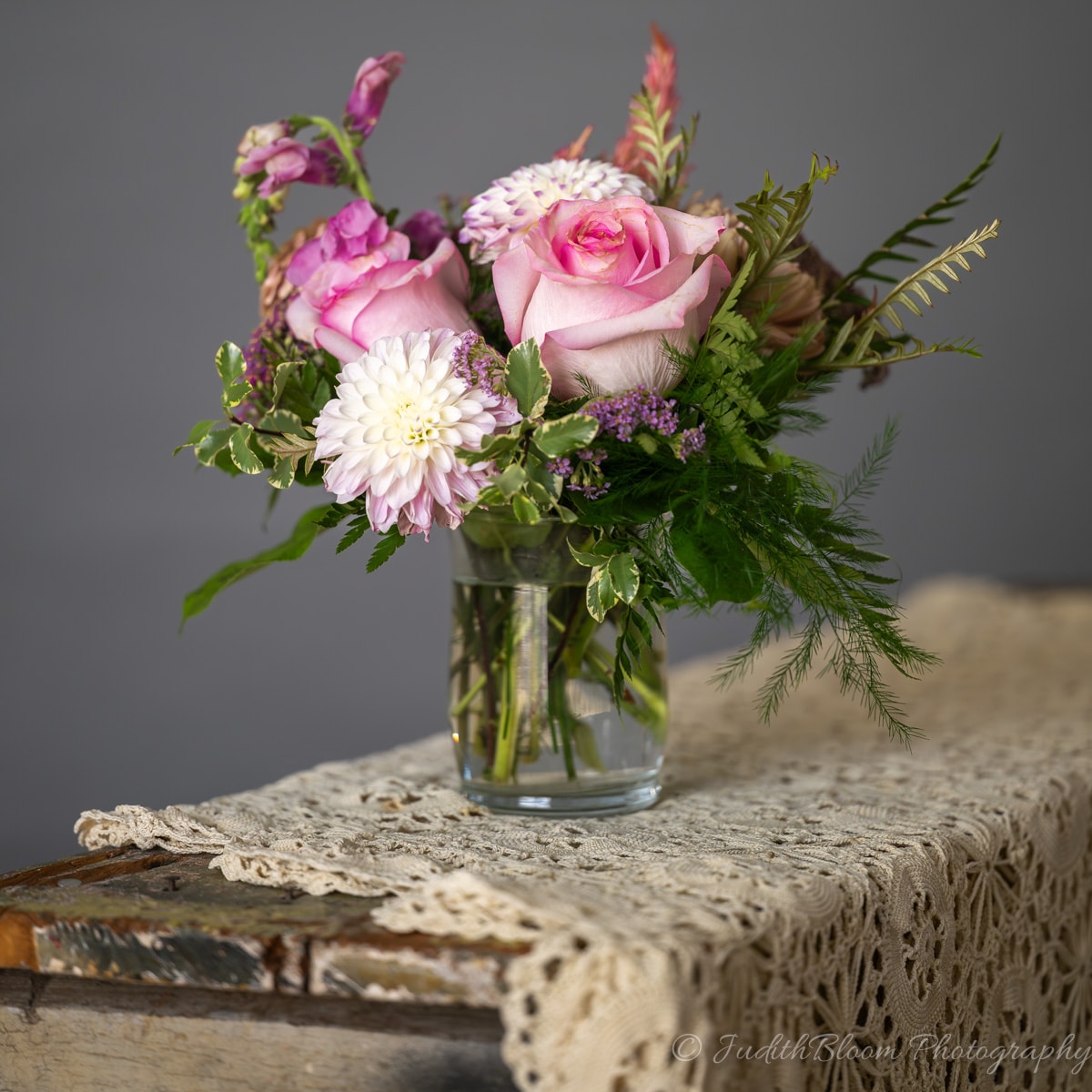 Pink roses and white dahlia in a glass jar on a lace-covered antique trunk